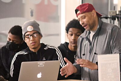 A man and several young men gather around a laptop, focused on the screen together
